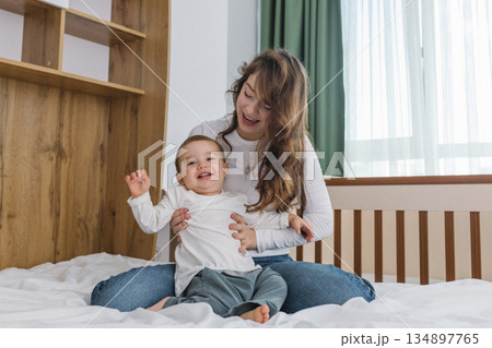 Young mother playing with her baby at home, enjoying a warm and cozy family moment full of tenderness, comfort, natural light, and playful parent-child connection. 134897765