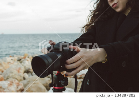 Female photographer adjusting professional camera on tripod outdoors by the sea, focusing the lens while preparing for a creative photo shoot on a rocky coastline. 134897802
