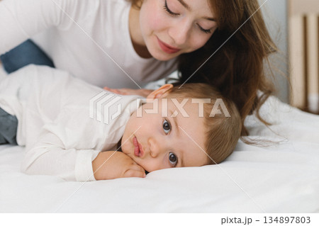 Cozy moment of a young woman and baby relaxing together at home, lying on a soft bed and smiling in natural light. Cozy moment of a young woman and baby relaxing together at home, lying on a soft bed and smiling in natural light. 134897803