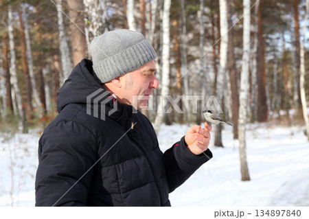 A man stands in a snowy forest. He is feeding a small bird from his hand. 134897840