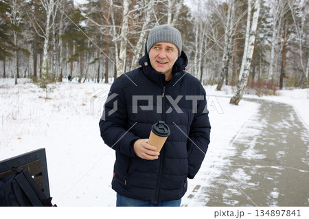 A happy man walks through a snowy forest in winter, holding a cup of hot drink in his hands A happy man walks through a snowy forest in winter, holding a cup of hot drink in his hands 134897841