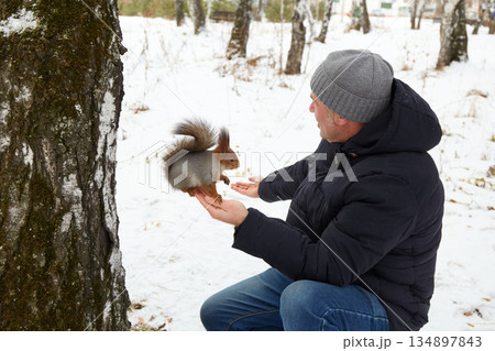 A man sits by a tree in a snowy forest, hand-feeding a squirrel. 134897843