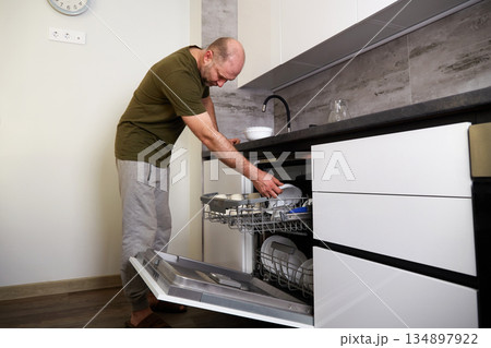 Man loading dishes into a dishwasher in a modern kitchen while standing on a wooden floor 134897922