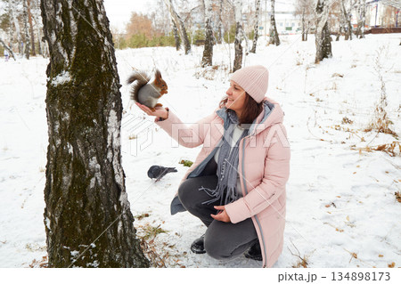 A woman sits by a tree in a snowy forest. She reaches out to feed a squirrel some nuts. The squirrel lands on her hand. A woman sits by a tree in a snowy forest. She reaches out to feed a squirrel some nuts. The squirrel lands on her hand. 134898173