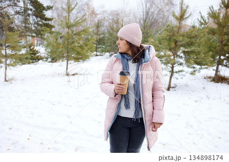 Woman walking on a snowy path in winter with a cup of drink in a forest setting 134898174
