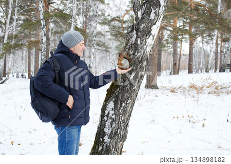 A man stands near a tree in a snowy forest. He holds out his hand to feed a squirrel. . A man stands near a tree in a snowy forest. He holds out his hand to feed a squirrel. . 134898182