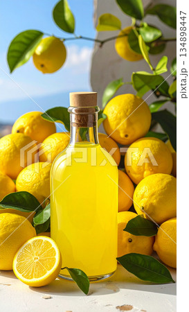 A bottle of bright yellow limoncello sits next to a pile of fresh lemons and leaves. 134898447