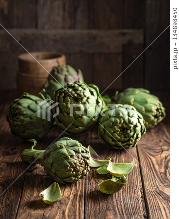 A rustic wooden table holds a pile of raw, green artichokes prepared in the traditional roman style. 134898450