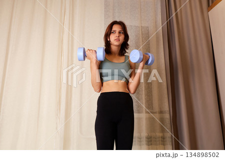 Young woman in sportswear is lifting dumbbells while standing in a home gym. . Young woman in sportswear is lifting dumbbells while standing in a home gym. . 134898502
