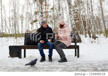 A man and woman are relaxing in a snowy winter park. They are sitting on a bench, drinking hot tea from a thermos A man and woman are relaxing in a snowy winter park. They are sitting on a bench, drinking hot tea from a thermos 134898516