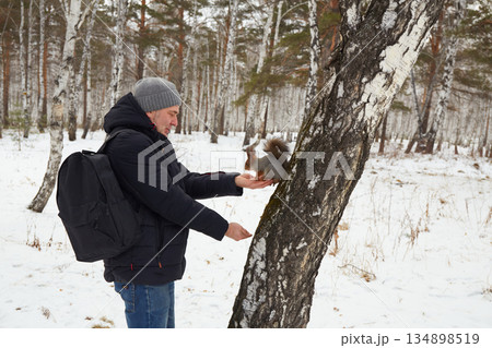 Man feeds squirrel while standing by tree in snowy forest during winter 134898519