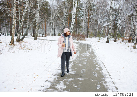 Woman walking on a snowy path in winter with a cup of drink in a forest setting 134898521
