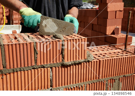 A skilled bricklayer is carefully applying mortar to red bricks, aligning them precisely to build a wall at a construction site 134898546
