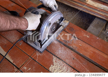 A construction worker uses a circular saw to cut wooden planks in a workshop 134899477