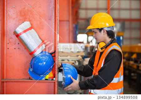 Industrial worker in safety vest and helmet placing blue hard hat on storage shelf inside factory. Concept of workplace organization, safety compliance and proper PPE management in industry. 134899508