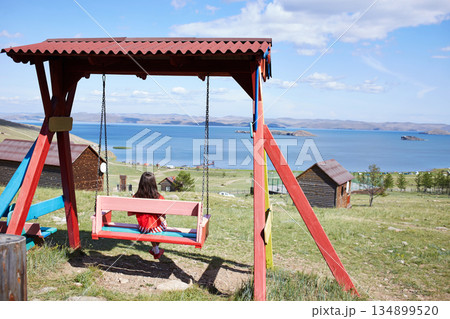 A young girl in a red shirt swings on a colorful swing set with a picturesque lake in the background. A young girl in a red shirt swings on a colorful swing set with a picturesque lake in the background. 134899520