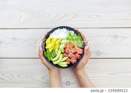 Poke bowl of salmon, rice, avocado, cucumber, mango, herbs in a woman's hands. Poke bowl of salmon, rice, avocado, cucumber, mango, herbs in a woman's hands. 134899522