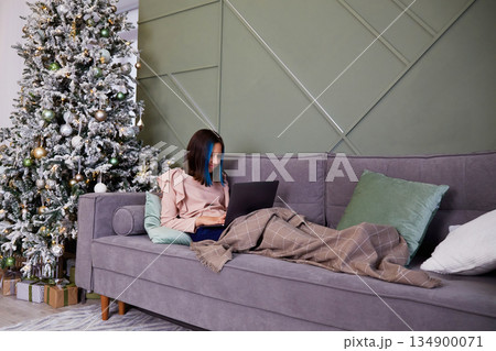 A pretty girl is working on a laptop, covered with a blanket. Christmas tree with toys and gifts. A pretty girl is working on a laptop, covered with a blanket. Christmas tree with toys and gifts. 134900071
