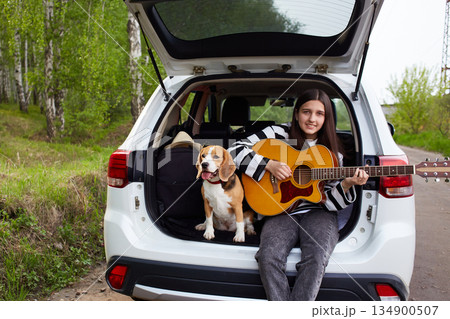 Young girl with guitar and beagle dog sitting in car trunk. Summer vacation stuff nearby. Young girl with guitar and beagle dog sitting in car trunk. Summer vacation stuff nearby. 134900507