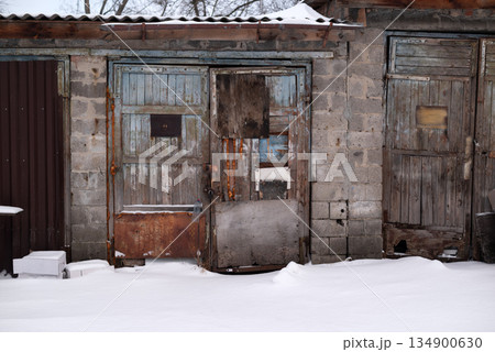 Weathered wooden garage doors in winter. Old abandoned shed with peeling paint and rusty metal patches covered in snow. Weathered wooden garage doors in winter. Old abandoned shed with peeling paint and rusty metal patches covered in snow. 134900630