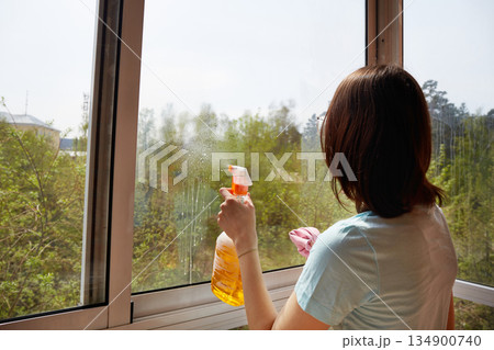 A young women is cleaning a large window with a squeegee, revealing a scenic outdoor view. 134900740