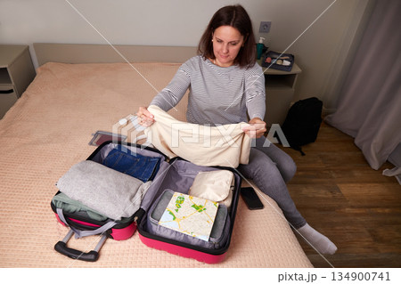 A woman is sitting on a bed, packing a suitcase for an upcoming trip, folding clothes and travel supplies A woman is sitting on a bed, packing a suitcase for an upcoming trip, folding clothes and travel supplies 134900741