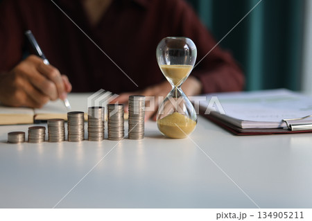 Rising stack of coins and running sand clock on the table. Growing of income, saving or return investment through time. Time and money concept. 134905211