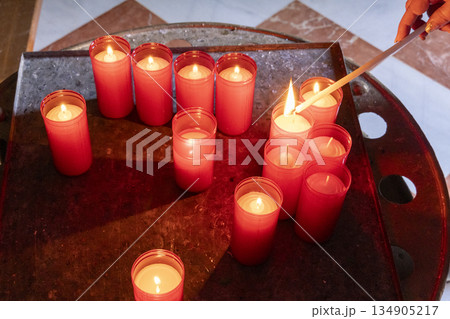 a female hand with a candle lighting a group of red candles as an offering inside a religious temple a female hand with a candle lighting a group of red candles as an offering inside a religious temple 134905217