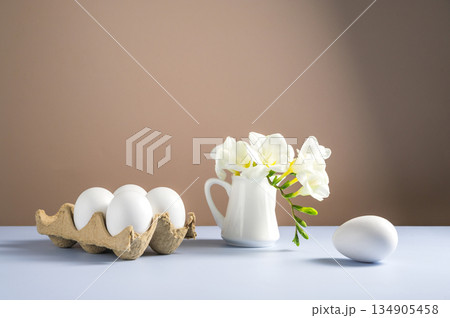 Easter eggs and flowers. White eggs in cardboard box and Freesia white spring bouquet in a ceramic jug 134905458