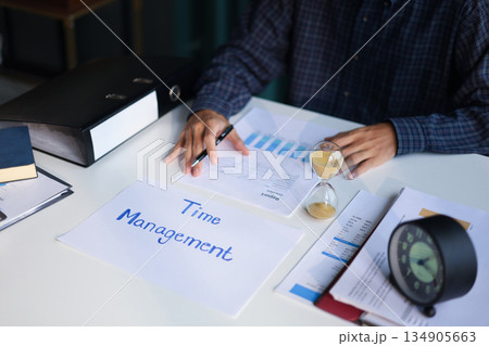 Time management concept. Businessman working on office desk with stack of documents, running sand clock and clock on the table. 134905663