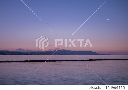 Lavender dusk over calm sea with faint moon above mountains 134906536