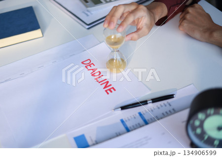 Male employee sitting on office desk holding running sand clock and Deadline paper on the table. Time management concept. 134906599