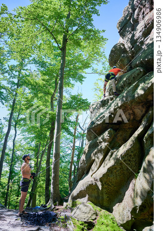 Male rock climber ascending rugged limestone cliff with harness and rope for safety. Sportsman climbing on vertical large boulder at Dobvush Rocks in Carpathian mountains, Ukraine. 134906986