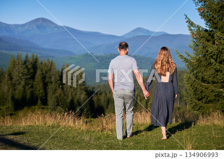 Couple holds hands, walk barefoot through meadow, facing stunning mountain view. Lush green forest and distant mountains under clear blue sky create serene and romantic atmosphere. 134906993