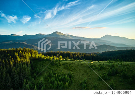 Aerial view of yellow tourist tent on sunlit, grassy plateau, surrounded by dense forest. Expansive scene reveals rolling green hills and majestic mountains, creating idyllic and serene camping spot. 134906994