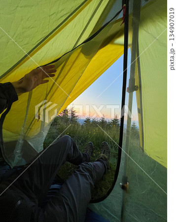 Camper peeks out from yellow tent at dawn, revealing serene view of grassy terrain and distant trees. Dewdrops glisten on tent fabric, with hiking boots placed at tent's entrance. Camper peeks out from yellow tent at dawn, revealing serene view of grassy terrain and distant trees. Dewdrops glisten on tent fabric, with hiking boots placed at tent's entrance. 134907019