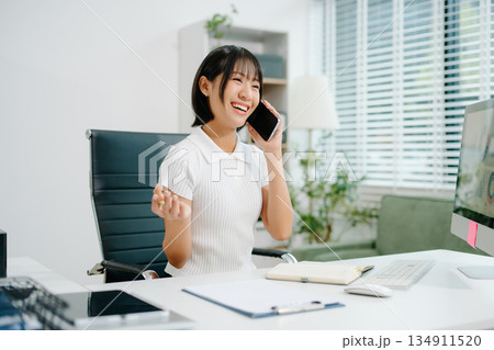 Asian Business woman Talking on the phone and using a laptop with a smile while sitting at office Asian Business woman Talking on the phone and using a laptop with a smile while sitting at office 134911520