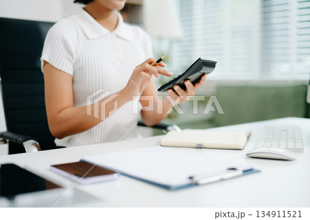 A young office woman calculating finances with a calculator and notebook at her desk, representing accounting, budgeting, financial planning, and office productivity. 134911521