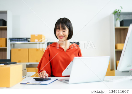 Young Asian woman using calculator and laptop in modern office with boxes. Ideal for themes like e-commerce, small business, shipping, startup Young Asian woman using calculator and laptop in modern office with boxes. Ideal for themes like e-commerce, small business, shipping, startup 134911607