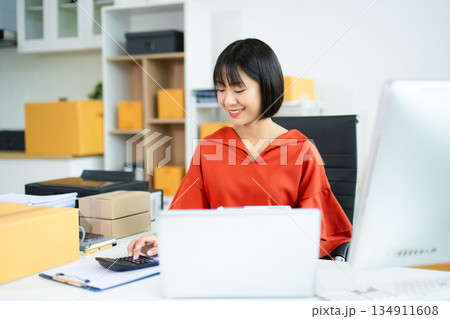 Young Asian woman using calculator and laptop in modern office with boxes. Ideal for themes like e-commerce, small business, shipping, startup Young Asian woman using calculator and laptop in modern office with boxes. Ideal for themes like e-commerce, small business, shipping, startup 134911608