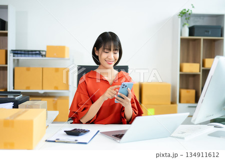 Young Asian woman using tablet in e-commerce office with boxes, laptop, and smile. Perfect for business, shipping, startup, and online seller concepts. Young Asian woman using tablet in e-commerce office with boxes, laptop, and smile. Perfect for business, shipping, startup, and online seller concepts. 134911612