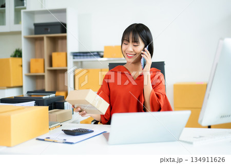 Smiling Asian entrepreneur talks on phone and holds package in modern office. Smiling Asian entrepreneur talks on phone and holds package in modern office. 134911626