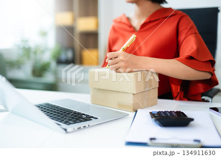 Young Asian entrepreneur writing on box in SME office with laptop and clipboard. Ideal for ecommerce, shipping, small business, logistics, and real people concepts. 134911630
