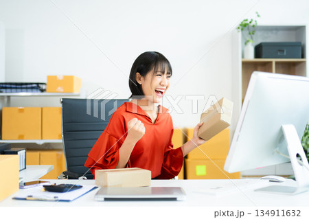 Smiling Asian woman raises fist in joy while holding smartphone. Perfect for business success, ecommerce wins, startup celebration, and real people themes. Smiling Asian woman raises fist in joy while holding smartphone. Perfect for business success, ecommerce wins, startup celebration, and real people themes. 134911632