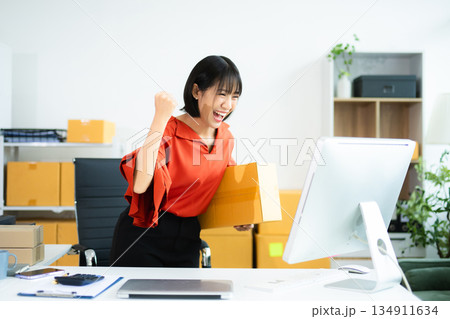 Smiling Asian woman raises fist in joy while holding smartphone. Perfect for business success, ecommerce wins, startup celebration, and real people themes. Smiling Asian woman raises fist in joy while holding smartphone. Perfect for business success, ecommerce wins, startup celebration, and real people themes. 134911634
