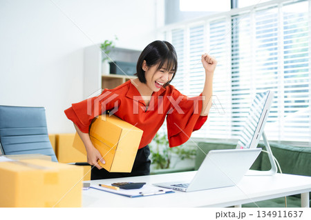 Smiling Asian woman raises fist in joy while holding smartphone. Perfect for business success, ecommerce wins, startup celebration, and real people themes. Smiling Asian woman raises fist in joy while holding smartphone. Perfect for business success, ecommerce wins, startup celebration, and real people themes. 134911637