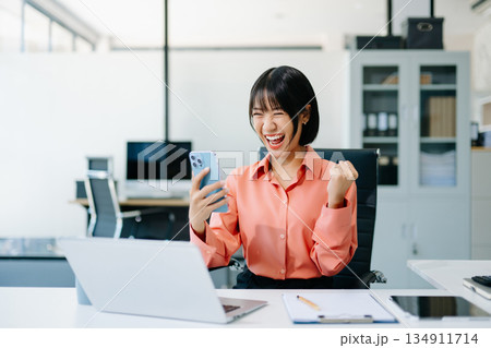 Happy Asian businesswoman celebrating success with smartphone at office desk. Concept of achievement, motivation, joy, and professional victory. 134911714