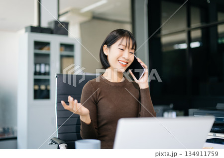 Asian Businesswoman Talking on the phone and using a laptop with a smile while sitting at office Asian Businesswoman Talking on the phone and using a laptop with a smile while sitting at office 134911759