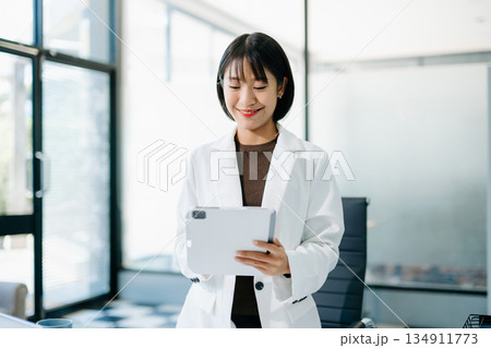 Smiling Asian woman in white blazer holding coffee cup and clipboard in bright office. Confident, stylish businesswoman balancing work and lifestyle. 134911773