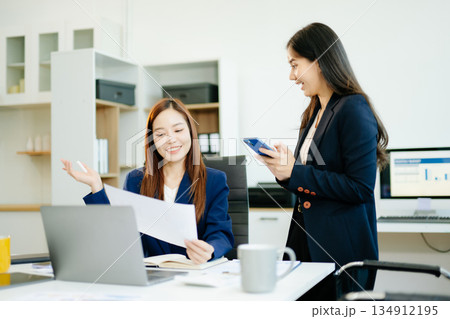 Two Asian businesswomen in suits analyze reports together at a modern office desk. A scene of teamwork, leadership Two Asian businesswomen in suits analyze reports together at a modern office desk. A scene of teamwork, leadership 134912195